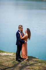 Cheerful newlyweds go holding hands and laughing, against the background of a lake and a green meadow. Cheerful groom and beautiful bride with curly hair walk in the meadow