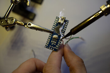 The process of soldering wires to an arduino nano board. The clamp holds the board. There is rosin smoke above the board. Hand holds solder. Soldering iron with a fireproof tip.