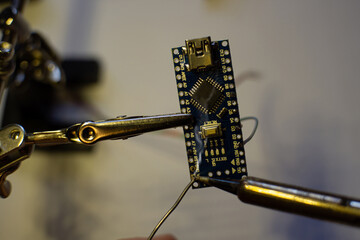 The process of soldering wires to an arduino nano board. The clamp holds the board, the hand holds the solder. The soldering iron melts the solder.