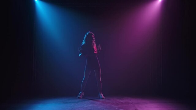 A slender long legged young female dancer who practices jazz funk movements in a dark studio is silhouetted. The girl enjoys dancing in the rays of beautiful light. Slow motion.