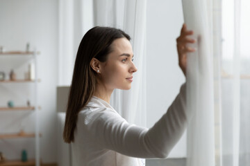 Young Caucasian woman open curtains in living room look in window distance thinking or dreaming. Millennial girl renter or tenant welcome meet new day in own home. Real estate, rental concept.
