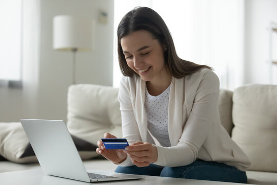 Smiling millennial Caucasian girl sit on couch at home shopping online on laptop with credit card. Happy young woman client buyer make web payment using secure internet service system on computer.
