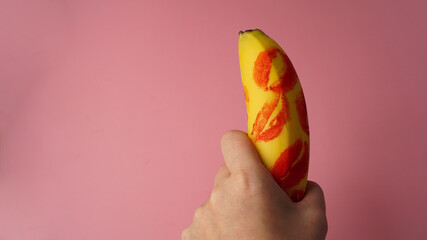Woman hand holding banana with red lipstick markings on a pink background