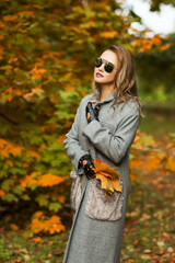 Fototapeta na wymiar Outdoors lifestyle fashion portrait of pretty young woman walking on the autumn park. Holding maple leaves. Wearing stylish grey coat, sunglasses and glovelettes. Enjoying autumn nature. Autumn colors