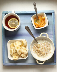 healthy breakfast on blue tray - porridge, cut banana, orange jam, lemon tea with spoon and tea strainer