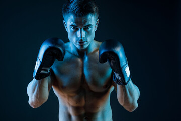 Muscular model sports young man in boxing gloves on dark background. Sexy torso. Male flexing his muscles.