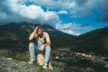 Young man grimaces, sitting on stone on background of village in mountains. Tired male tourist aping, resting on hill after active trekking in mountains.