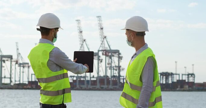 Engineers in special clothes and with masks on their faces work in quarantine at the seaport. Port workers work with a tablet and point to the side of the cranes.