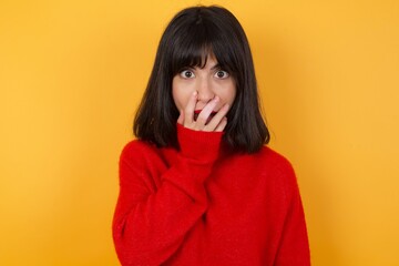 Emotional Caucasian brunette woman wearing red casual sweater isolated over yellow background gasps from astonishment, covers opened mouth with palm, looks shocked at camera. Reaction concept.