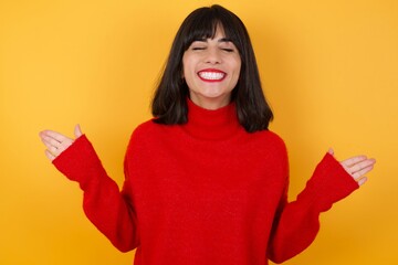 Emotive Caucasian brunette woman wearing red casual sweater isolated over yellow background laughs loudly, hears funny joke or story, raises palms with satisfaction, being overjoyed, 