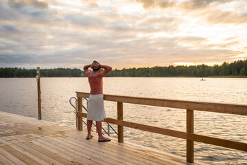 Sunset on the lake. Wooden bath or sauna with descent into the water. A man in a special hat for a bath is wrapped in a towel. He looks at the sunset and happily raises his hands up.