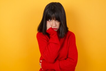 Very bored Caucasian brunette woman wearing red casual sweater isolated over yellow background holding hand on cheek while support it with another crossed hand, looking tired and sick,