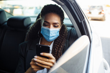 Fototapeta na wymiar Woman wearing a medical mask on a business travel around the city checking her mobile phone, chatting and calling. Business trips during pandemic, new normal and coronavirus travel safety concept.