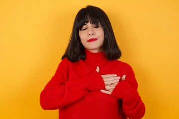 Caucasian brunette woman wearing red casual sweater isolated over yellow background smiling with hands on chest with closed eyes and grateful gesture on face. Health concept.