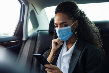 Young business woman in a mask checking her mobile cell phone on a backseat of a taxi during covid-19 quarantine. Business trips during pandemic, new normal and coronavirus travel safety concept.