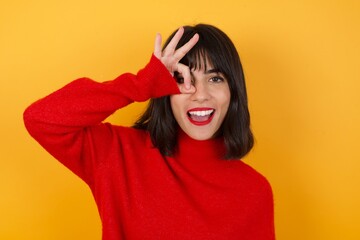 Caucasian brunette woman wearing red casual sweater isolated over yellow background doing ok gesture shocked with surprised face, eye looking through fingers. Unbelieving expression.