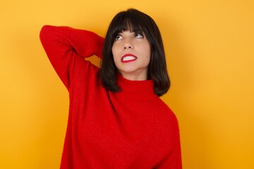 Caucasian brunette woman wearing red casual sweater isolated over yellow background confuse and wonder about question. Uncertain with doubt, thinking with hand on head. Pensive concept.