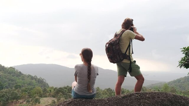 Rear view of two hikers with backpacks on the background of mountains. A woman is sitting on a hill. A man takes a landscape picture on camera	