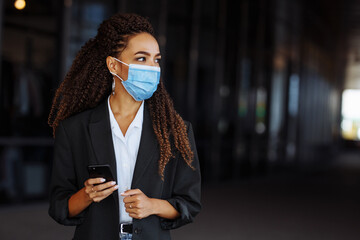 Young businesswoman wearing a medical mask stands near the office center. Officially looking girl with a phone in her hands waiting outside. Leading business during Covid-19 pandemic concept.