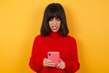 Focused Caucasian brunette woman wearing red casual sweater isolated over yellow background use smartphone reading social media news, or important e-mail