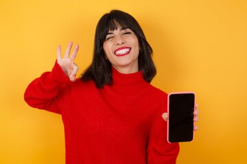 Caucasian brunette woman wearing red casual sweater isolated over yellow background holding in...