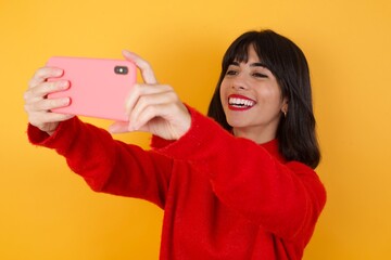 Caucasian brunette woman wearing red casual sweater isolated over yellow background taking a selfie...