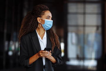 Young businesswoman wearing a medical mask stands near the office center. Officially looking girl with a phone in her hands waiting outside. Leading business during Covid-19 pandemic concept.