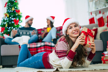 Beautiful young happy smiling woman in a Christmas sweater and Santa hat is holding a colorful gift box in hands at home on the background of her friends and Christmas tree.
