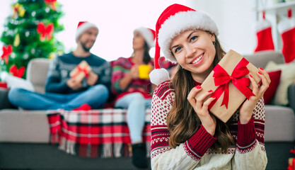 Close up photo of beautiful young happy smiling woman in a Christmas sweater and Santa hat is holding a gift box in hands at home on the background of her friends and Christmas tree.
