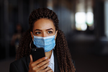 Young businesswoman wearing a medical mask stands near the office center. Officially looking girl with a phone in her hands waiting outside. Leading business during Covid-19 pandemic concept.