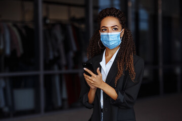 Young businesswoman wearing a medical mask stands near the office center. Officially looking girl with a phone in her hands waiting outside. Leading business during Covid-19 pandemic concept.