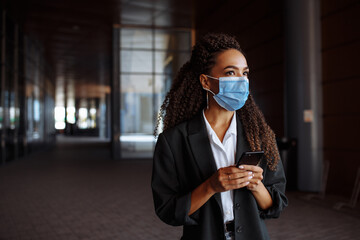 Young businesswoman wearing a medical mask stands near the office center. Officially looking girl with a phone in her hands waiting outside. Leading business during Covid-19 pandemic concept.