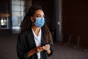 Young businesswoman wearing a medical mask stands near the office center. Officially looking girl with a phone in her hands waiting outside. Leading business during Covid-19 pandemic concept.