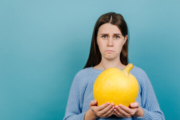 Tired unhappy young woman feels disgust, holds small ripe yellow pumpkin, has dissatisfied facial expression, dressed in knitted sweater, isolated over pastel blue studio background. Autumn concept