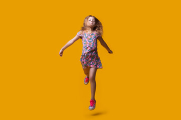 Cute blonde lady running on a yellow studio wall wearing a summer dress and gesturing a hurry