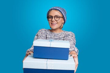 Caucasian woman with hat and glasses is givving some presents at camera smiling on a blue studio wall