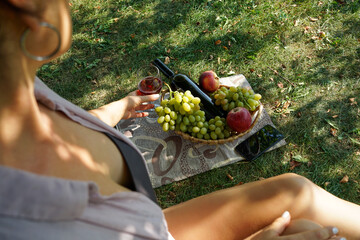 A girl with a smartphone on a picnic by the lake sits on a green lawn. She is wearing casual clothes: a light shirt and denim shorts. Nearby is a fruit basket and a bottle of wine