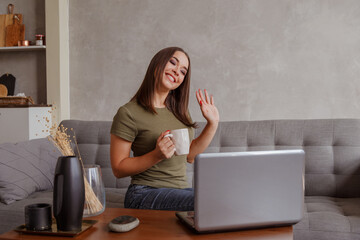 Portrait of pleasant young woman with mug looking at camera, waving hello. Pretty millennial lady communicating with parents via video call
