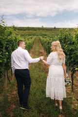 Fototapeta na wymiar Groom and bride walking in vineyard in their wedding day. Blond hair woman in white dress hold wedding bouquet in hands