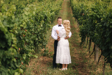 Groom and bride walking in vineyard in their wedding day. Blond hair woman in white dress hold wedding bouquet in hands