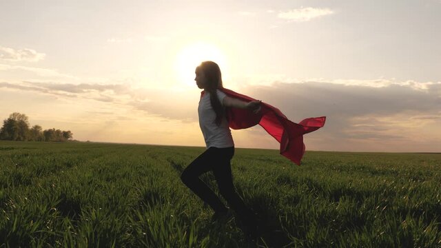 teenager dreams of becoming superhero. young girl in red cloak, dream expression. happy superhero girl, runs on green field in red cloak, cloak flutters in wind. child plays and dreams. Slow motion.