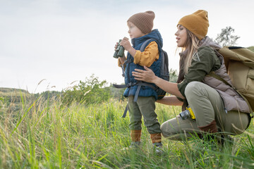 Young woman with backpack squatting by her little son with binoculars on trip