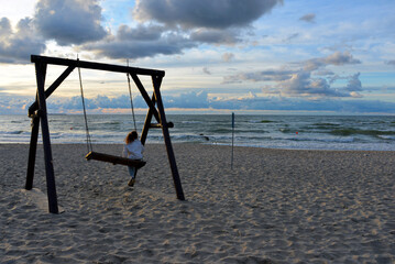 a little girl rides on a wooden swing against the background of the waves of the Baltic sea and the beautiful sky.