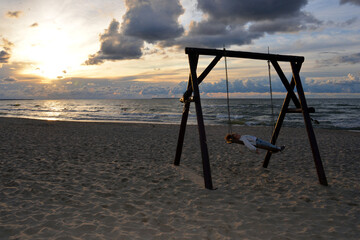 a little girl rides on a wooden swing against the background of the waves of the Baltic sea and the beautiful sky.