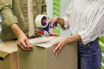 Close up of unrecognizable couple packing boxes with tape gun while standing in self storage unit,...