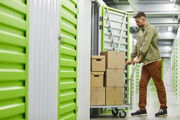 Full length side view at handsome bearded man loading cart with cardboard boxes into self storage...