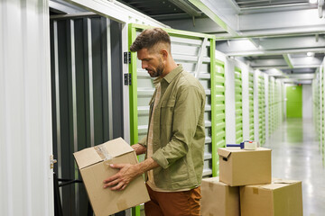 Side view at bearded adult man loading cardboard boxes into self storage unit, copy space