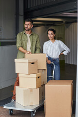 Vertical full length portrait of modern couple standing by cart with cardboard boxes while loading...