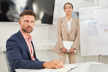 Portrait of handsome businessman looking at camera while sitting at table in conference room with female colleague or secretary in background, copy space