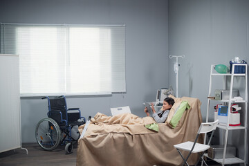Pretty, little girl with a tablet in a hospital bed, a wheelchair in the background.